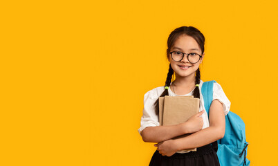 Asian Schoolgirl Embracing Book Standing In Studio On Yellow Background
