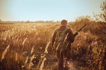 Young hunter in the sunny field with military backpack and shotgun waiting for flying ducks