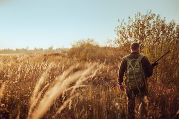 Young hunter in the sunny field with military backpack and shotgun waiting for flying ducks