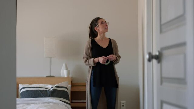 Stylish woman looks around room holding painters tape - ready to paint bedroom - wide shot
