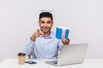 Congratulations to you! Joyful angelic businessman with nimbus on head holding gift box and pointing to camera, sitting at laptop workplace, greeting with professional holidays. studio shot isolated