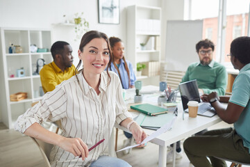 Portrait of smiling young woman looking at camera while sitting at table during meeting with creative business team in background, copy space