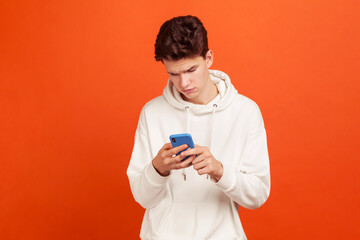 Serious concentrated teenager in casual sweatshirt with hood holding cellphone in hands, scrolling...