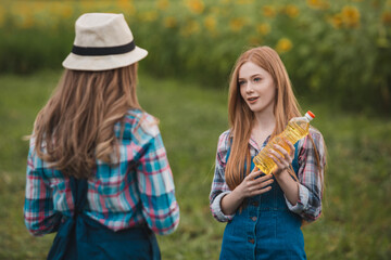 Two beautiful farmerettes having a discussion while one of them is holding a bottle of golden sunflower oil in the middle of a green blooming sunflower field.