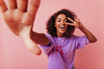 Blithesome black woman with wavy hair making selfie on rosy background. Wonderful girl with positive smile taking picture of herself.