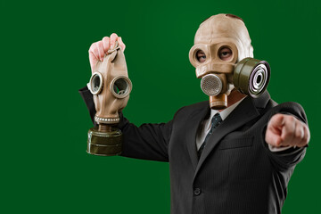 Portrait of a man wearing a dark business suit and a gas mask while holding a gas mask and pointing towards the camera, isolated on green background.