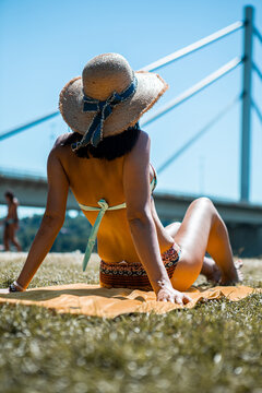 A back view of a young tanned woman sitting on a towel on the beach and looking in the direction of the bridge. She wears a colorful bikini and has a knitted hat on her head.