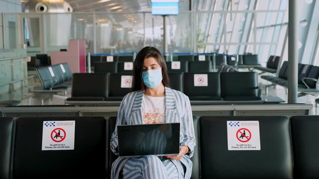 woman, in protective mask, uses laptop, while sitting in empty airport lounge, waiting to board a flight. air travel re-opened after coronavirus outbreak end. Opening borders. travel open.