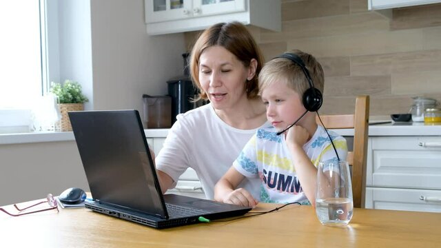 Little boy learning online together with his mom in the kitchen with computer and wearing headphone