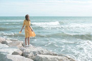 Young beautiful woman in a bright yellow dress stands on the pier on the seashore or ocean with her arms wide open. Concept of freedom, enjoyment of nature, thoughtfulness, mindfulness.