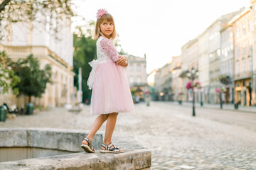 Fashion full length portrait of a cute little blond girl, wearing pink dress and flower in hair, posing to camera, while standing on ancient stone fountain in old European city