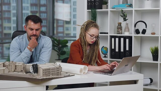 Charming serious smart stylish woman with dreadlocks working on computer while her high-skilled handsome attantive bearded male colleague working with wooden model of building in design office