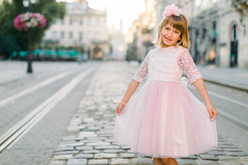 Outdoor portrait of a cute little blond Caucasian girl in a city, wearing beautiful elegant pink dress and flower in her hair. Summer city portrait. Copy space