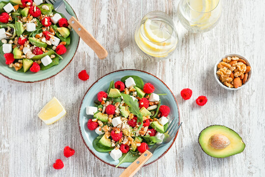 Light salad with raspberry, baby spinach, feta cheese, avocado and walnuts, top view. Fresh original summer salad with lemon water on wooden table top