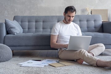 Man working on laptop computer sitting on the floor with his diary and official papers at home.