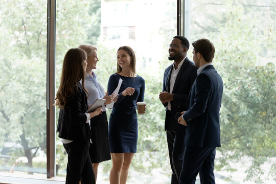 Five Successful Different Age And Ethnicity Office Employees Chatting Standing In Office Hall Against Panoramic Window Take Break Resting After Seminar, Wait Negotiations Start, Feels Prepared Concept