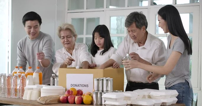 Group of asian volunteers family putting food products in donation box together as charity workers and members of community work to the poor during the Coronavirus pandemic. social distance concept.
