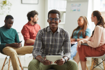 Multi-ethnic group of people sitting in circle while discussing business project in office, focus on young African-American man smiling at camera in foreground, copy space