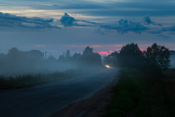 Russian countryside road with distant headlights of a car at summer foggy night