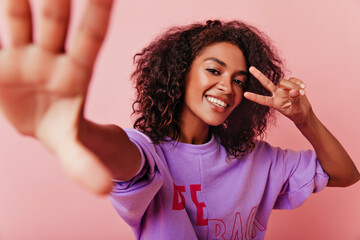 Appealing african woman making selfie with peace sign. Indoor photo of emotional laughing girl...