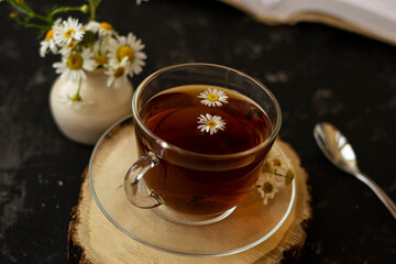 English tea with chamomile stands on a wooden frame and a teaspoon and an open book for reading lies on a black background. Nearby is a white vase with summer chamomile flowers