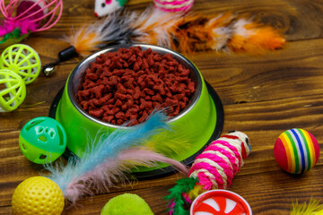 Set of toys for cat and bowl with dry pet food on wooden background. Pet care concept