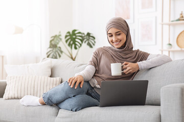 Home Passtime . Cheerful Arabic Woman Watching Movies On Laptop And Drinking Coffee