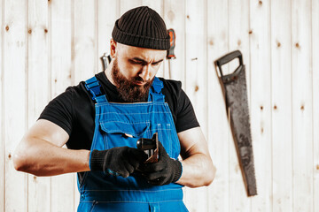 A dark-haired man with a beard and in overalls holds in hands and adjusts black jack plane,  in the background a lot of wooden boards. Work with wooden .