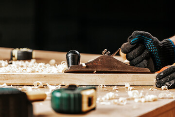 Close up of the an young  man builder in work clothers and black gloves handles a wooden bar with a  jack plane in the workshop