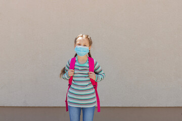 Girl wearing a protective mask with a backpack behind his back in the schoolyard on the first school day after isolation or vacation. Children are happy to go back to school