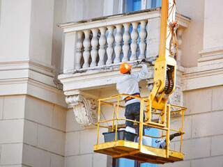 Worker repairing historic architecture of old facade. Plasterer in hardhat paint balcony of building at height in cradle. Renovation work. Construction worker working at height in lifting bucket