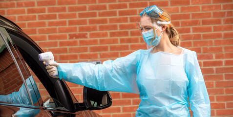 Salisbury, England, UK. August 2020. Woman Covid-19 tester wearing protective clothing, visor, mask and goggles using forehead thermometer to check temperature