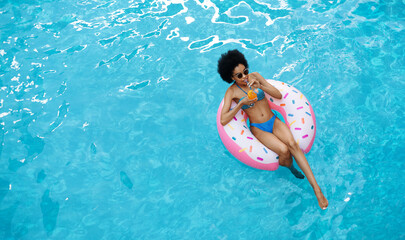 Relaxed black lady floating on inflatable ring and enjoying her cocktail drink at pool, top view. Space for text