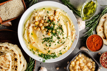 Hummus topped with chickpeas, olive oil and green coriander leaves on stone table with pita bread and spices aside. Flat lay