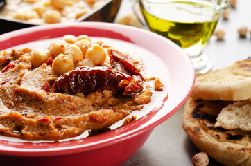 Hummus in clay dish topped with olive oil, chickpeas and sun dried tomatoes on stone table served with traditional Pita bread