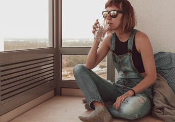 A disheveled girl in dark glasses sits on the balcony and drinks water from a glass. The concept of...