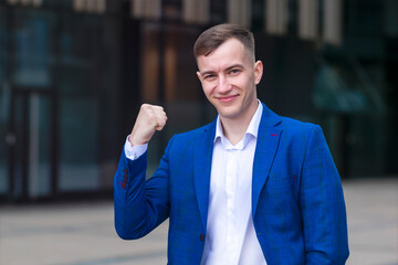 Portrait of handsome confident positive man, successful happy businessman in formal suit, jacket raise fist, celebrating success, victory, triumph.Young European blonde cheerful guy smiling outdoors