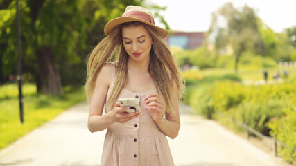 Young blonde woman wearing hat using phone in a city park.