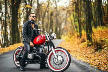 Bearded brutal man in sunglasses and leather jacket sitting on a motorcycle on the road in the forest