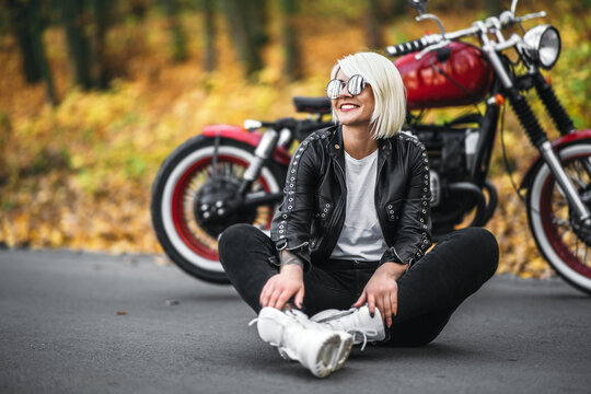 Pretty blonde biker girl in sunglasses sitting near red motorcycle on the road