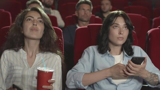 Medium shot of two multiethnic beautiful women sitting in cinema, using smartphones then putting them away and starting watching movie
