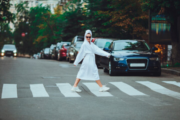 A glamorous lady in a white bathrobe with a white towel on her head and elegant sunglasses is walking along the street of a big city