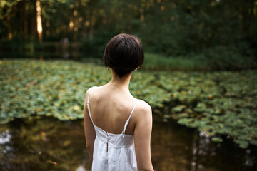 Naklejka premium Rear shot of unrecognizable short haired young woman in strap white dress relaxing by pond in park, enjoying beautiful landscape and hot summer day. Back view of female walking outdoors alone