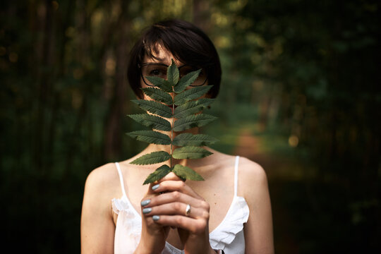 Wood nymph in summer white dress holding big green leaf, hiding her face from sunlight. Outdoor image of beautiful young woman spending nice time in forest alone, feeling deep connection to nature