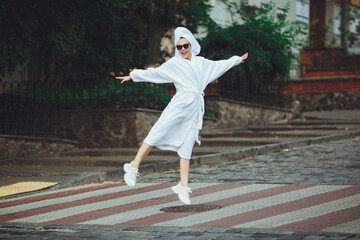 A glamorous lady in a white bathrobe with a white towel on her head and elegant sunglasses is walking along the street of a big city