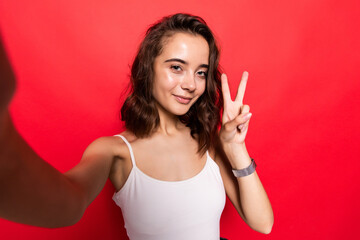 Young woman having fun and taking selfie with gesturing peace sign isolated over red background