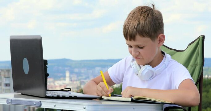 the boy sits working at a laptop and writes data from the monitor to a notebook and looks into a monocular telescope