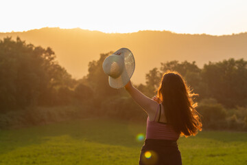 Young girl from the back holding a hat with her hand on a green meadow with sunset color
