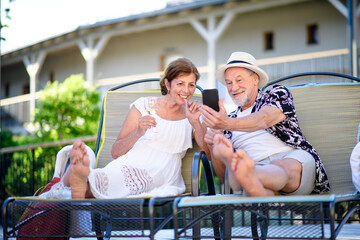 Senior couple with wine and smartphone outdoors on holiday, relaxing.