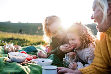 Small girl with mother and grandmother having picnic in nature at sunset.